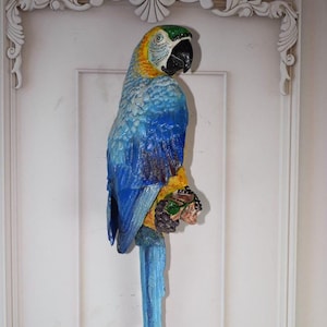 Op de afbeelding: Een blauwe en gele papegaai-figuur met een lange staart. De papegaai zit op een tak met groene bladeren. Het beeldje is gemaakt van geschilderd hout en heeft een realistisch uiterlijk.
