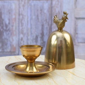 May include: A golden brass bell with a rooster finial and a matching candle holder on a round base. The bell and candle holder are on a marble-like surface, with a weathered white door in the background.