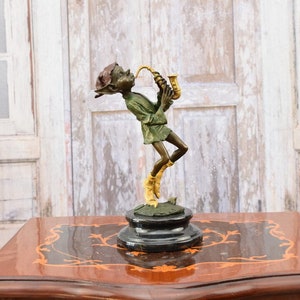 Peut inclure: Une sculpture en bronze d'un elfe jouant du saxophone. L'elfe porte une tunique verte et des bottes jaunes, debout sur une base ronde. Le saxophone est de couleur or. La sculpture est posée sur une table en bois.