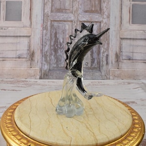 May include: A black and clear glass sculpture of a marlin leaping out of the water. The sculpture is on a round, marble-topped table with a gold base.