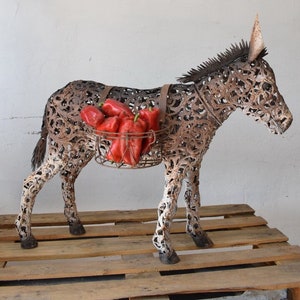 May include: A rustic metal donkey sculpture with a basket filled with red bell peppers. The donkey is made of intricate metalwork and stands on a wooden pallet. The sculpture has a brown and white color scheme.
