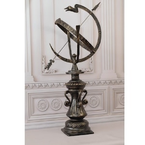 May include: A bronze-colored armillary sphere, a historical astronomical device, stands on a decorative pedestal. The sphere features intersecting rings, an arrow, and a bird-like figure at the top. The base has a square shape with ornate detailing, and the overall design is antique.