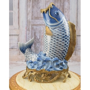 May include: A blue and white porcelain fish sculpture with gold accents. The fish is standing on a gold metal base with a decorative design. The fish is a traditional Chinese symbol of good luck and prosperity.