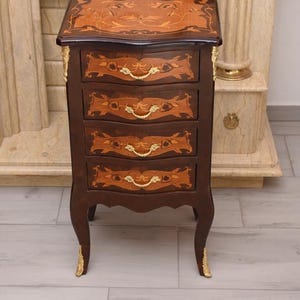 Può includere: Un comodino alto in legno scuro con quattro cassetti caratterizzati da un intricato intarsio floreale. Il mobile è arricchito da pomelli in metallo dorato e da decorazioni metalliche applicate sulle gambe curve.