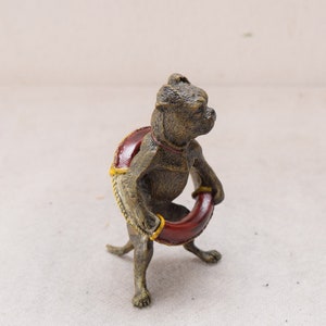 May include: A bronze-coloured figurine of a dog holding a red ring. The dog has a red and yellow saddle. The figurine is detailed with a textured finish, showcasing the dog's features and the ring it holds.