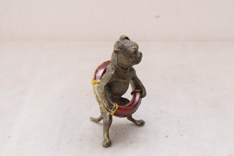 May include: A bronze-coloured figurine of a dog holding a red ring. The dog has a red and yellow saddle. The figurine is detailed with a textured finish, showcasing the dog's features and the ring it holds.