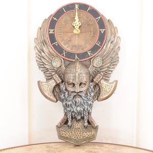 May include: A bronze-colored wall clock with a Viking theme. The clock face has a compass design and Roman numerals. The clock is adorned with a Viking helmet, axes, and wings. The clock measures approximately 12 inches in diameter.