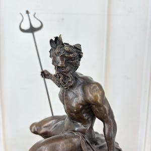 May include: A bronze sculpture of a seated male figure holding a trident. The figure has a beard and a crown of leaves. The sculpture is detailed and has a dark brown patina, suggesting an antique or vintage piece.