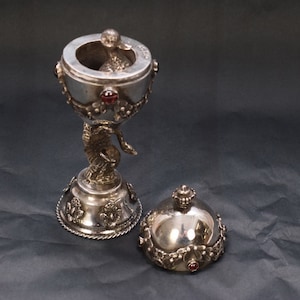 Pode incluir: Um cálice de metal prateado com tampa abobadada removível, apresentando uma haste central em forma de pássaro, detalhes florais em relevo e pequenas pedras vermelhas incrustadas na borda e na base, com uma figura de pássaro dentro da taça.