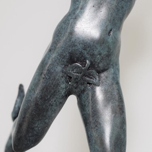 May include: A bronze-colored sculpture fragment depicting a human figure. The sculpture shows the torso and leg of a person, with a leaf detail. The sculpture has a weathered appearance.