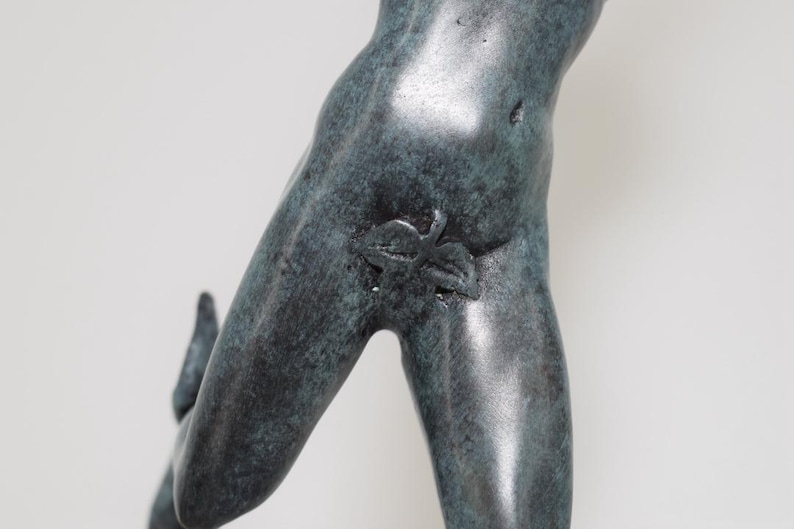 May include: A bronze-colored sculpture fragment depicting a human figure. The sculpture shows the torso and leg of a person, with a leaf detail. The sculpture has a weathered appearance.
