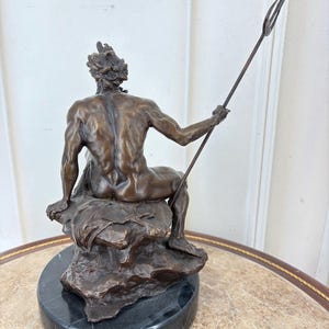 May include: A bronze sculpture of a seated figure holding a trident. The figure is seated on a rock formation atop a black marble base. The sculpture has a detailed, aged appearance.