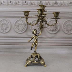 Könnte beinhalten: Ein goldfarbener Kerzenleuchter aus Metall mit einer Cherubfigur, die eine Kerze hält. Der Kerzenleuchter hat vier Arme und eine runde Basis.