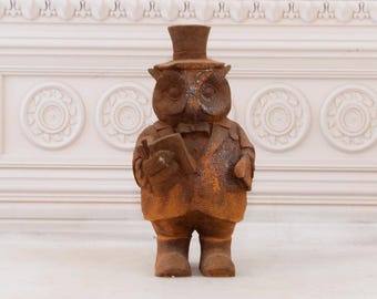 May include: A rusty metal owl statue wearing a top hat and holding a book. The owl is standing on a white surface.