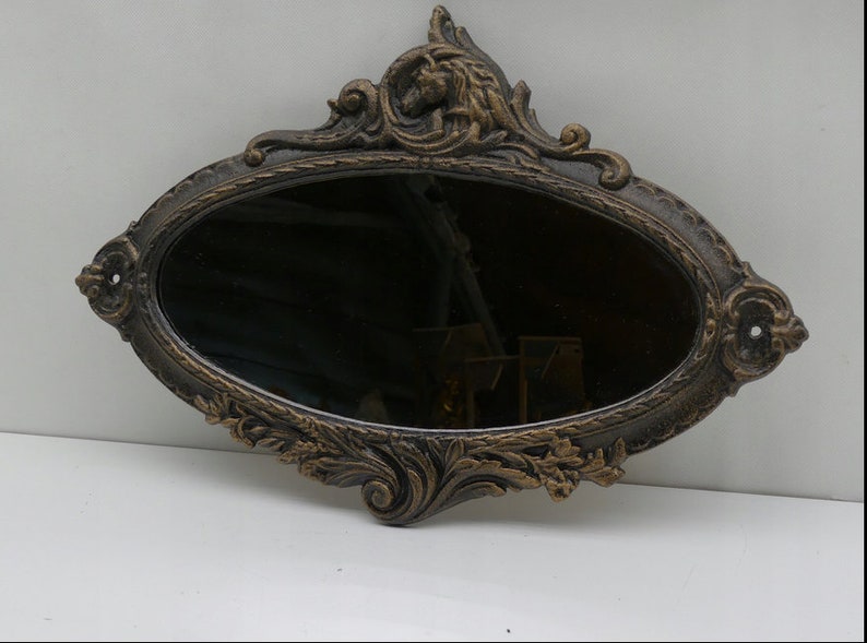 May include: An oval-shaped mirror with an ornate, gold-painted, cast iron frame. The frame features intricate floral and scrollwork designs.