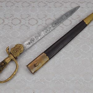 Op de afbeelding: Een jachtdolk met een getextureerd bruin handvat, een goudkleurige metalen stootplaat en een puntig stalen lemmet met gedetailleerde gravures van wilde dieren. De dolk wordt vergezeld door een zwarte schede met goudkleurige metalen beslag.