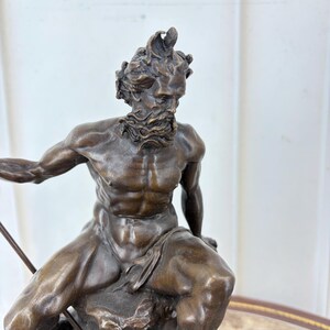 May include: Bronze sculpture of a muscular male figure, seated on a rock, holding a spear. The figure has a beard and a crown-like headpiece. The sculpture is a dark bronze color and is set on a black base.