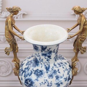 May include: A large white porcelain vase with a blue floral pattern and two gold-toned handles shaped like women holding the vase.
