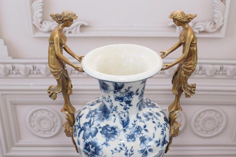 May include: A large white porcelain vase with a blue floral pattern and two gold-toned handles shaped like women holding the vase.
