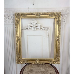 May include: A rectangular gold-coloured picture frame featuring ornate, raised relief detailing along the edges and corners. The frame has a hollow centre and displays intricate scrollwork and floral-inspired patterns throughout its metallic surface.