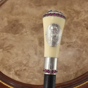 Op de afbeelding: Antieke wandelstok met een crèmekleurige handgreep versierd met een zilveren medaillon en een band van rode edelstenen. De handgreep is afgewerkt met meer rode edelstenen. De schacht is zwart.