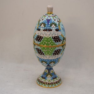 Op de afbeelding: Een decoratieve eivormige container met een deksel, gemaakt van metaal en versierd met kleurrijk email. De container staat op een standaard met een vergelijkbaar ontwerp. Het ei is blauw, groen, wit en zwart met gouden accenten.