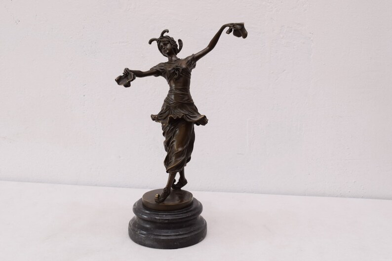 Op de afbeelding: Bronzen beeld van een dansende vrouw, die in elke hand een paar schoenen vasthoudt. Het beeld staat op een zwarte marmeren sokkel.