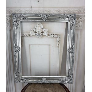 May include: A rectangular silver picture frame featuring ornate, raised floral and scrollwork detailing along the edges and corners. The frame has a metallic finish with a textured border and a smooth inner edge, designed for displaying artwork or mirrors.