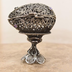Puede incluir: Elegante joyero en forma de huevo, plateado, con intrincados detalles de filigrana y pequeñas gemas moradas. La tapa con bisagras revela un interior vacío. Se apoya sobre una base decorativa con diseño floral.
