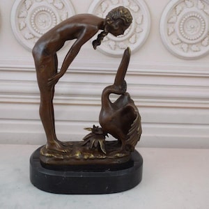 Könnte beinhalten: Bronzeskulptur einer Frau, die auf einen Pelikan blickt. Die Skulptur steht auf einem schwarzen Marmorsockel.