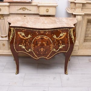 May include: A dark brown antique chest of drawers with a marble top and ornate gold accents. The chest features curved legs and detailed floral inlay designs. The furniture is set against a neutral background.