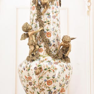 May include: A decorative ceramic vase with a floral pattern in orange, blue, and purple. The vase features three bronze cherub figures and a bronze floral garland. The base and rim are also bronze.