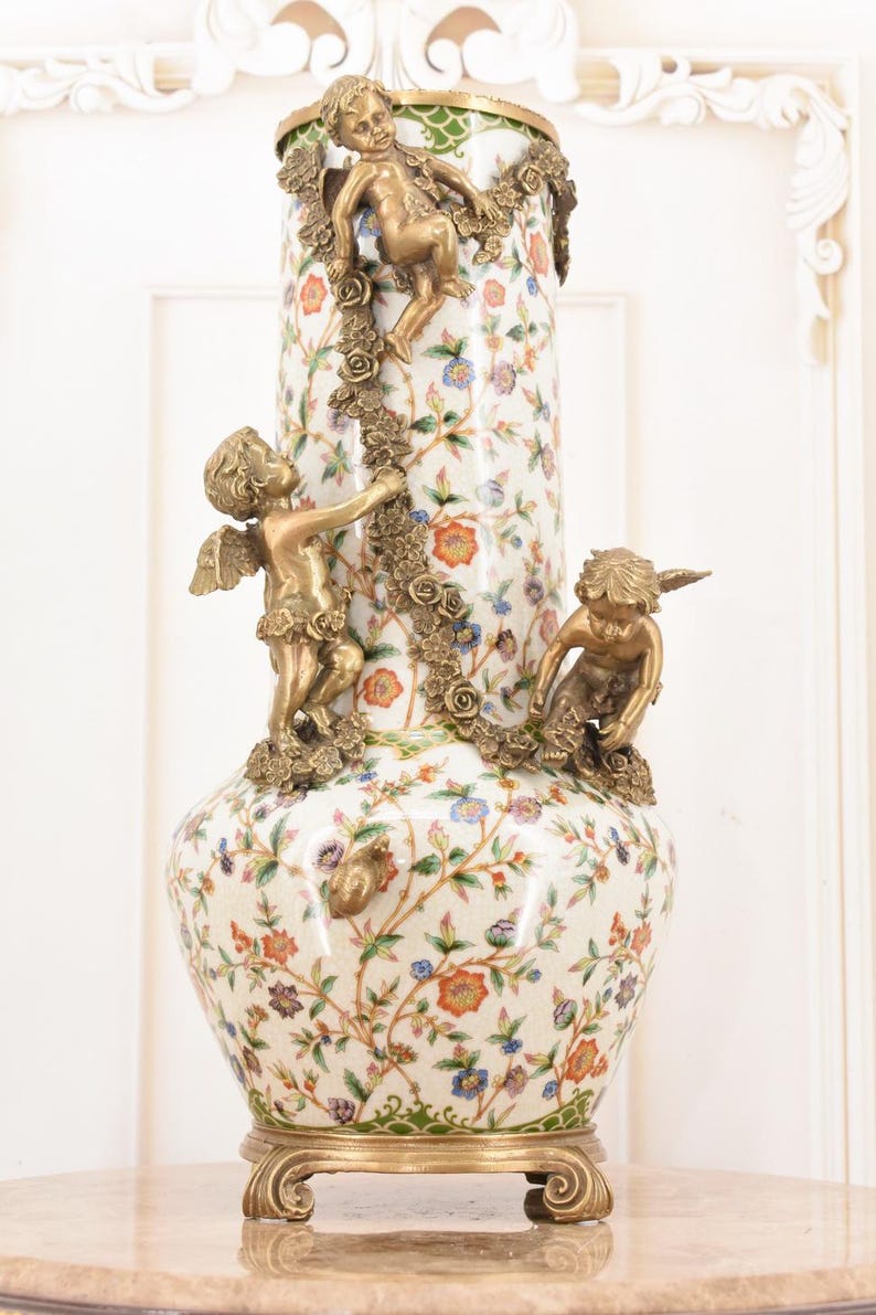 May include: A decorative ceramic vase with a floral pattern in orange, blue, and purple. The vase features three bronze cherub figures and a bronze floral garland. The base and rim are also bronze.