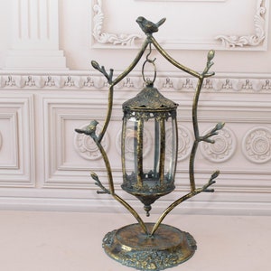 Può includere: Una lanterna in metallo in stile antico con un uccello in cima. La lanterna ha un involucro di vetro trasparente ed è sostenuta da una cornice decorativa con accenti a forma di ramo. La base è rotonda e ornata, con una finitura oro e verde acqua anticata.