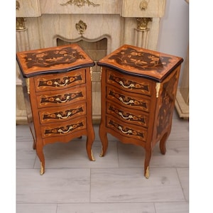 Puede incluir: Dos mesitas de noche de madera a juego con intrincados diseños de incrustaciones florales en las tapas y frentes de los cajones. Las mesitas de noche tienen patas curvas con detalles dorados y tiradores ornamentados. La madera tiene un tono marrón rojizo cálido.