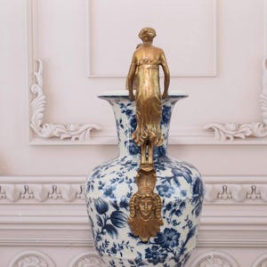 May include: A large white and blue porcelain vase with a gold-toned metal figure on top. The vase has a floral pattern and is decorated with gold accents.