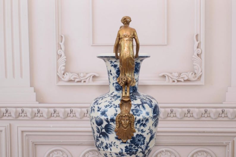 May include: A large white and blue porcelain vase with a gold-toned metal figure on top. The vase has a floral pattern and is decorated with gold accents.
