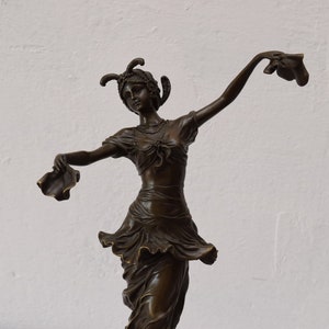 Op de afbeelding: Een bronzen beeld van een vrouw in een vloeiende jurk, die in elke hand een stuk stof vasthoudt. Ze draagt een hoofdtooi met veren en een ketting. Het beeld staat op een witte achtergrond.