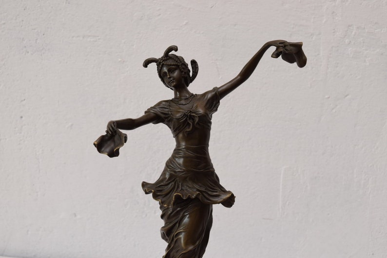 Op de afbeelding: Een bronzen beeld van een vrouw in een vloeiende jurk, die in elke hand een stuk stof vasthoudt. Ze draagt een hoofdtooi met veren en een ketting. Het beeld staat op een witte achtergrond.
