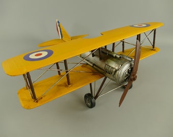 May include: A yellow biplane model featuring a silver engine, a brown propeller, and roundels on the wings. The aircraft has brown struts, wire bracing, and black wheels. The tail fin displays red, white, and blue stripes, reflecting a classic design.
