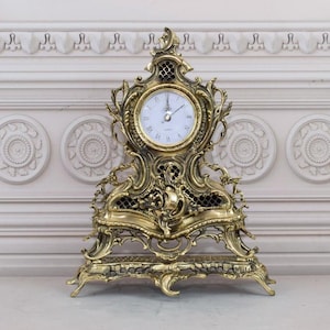 May include: An ornate, decorative clock with a gold-toned finish. The clock has a white face with black hands and a detailed, intricate design with a base that resembles a pedestal.