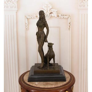 May include: A bronze sculpture of a woman and a dog. The woman is wearing a detailed top and a short skirt. The dog is sitting at her feet. The sculpture is on a black base and a round table.