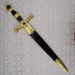 以下が含まれることがあります： 黒いハンドルと鞘を備えた装飾用の短剣で、金色の金属製の装飾が施されています。ヒルトには精巧な彫刻が施されたクロスガードがあり、鞘の先端も同様の金色の金属パーツで仕上げられています。