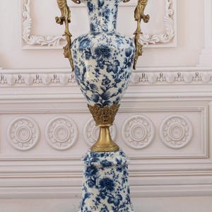 May include: A large, white porcelain vase with a blue floral pattern and gold accents. The vase has two gold handles in the shape of women holding up the vase. The vase is on a square base with gold feet.