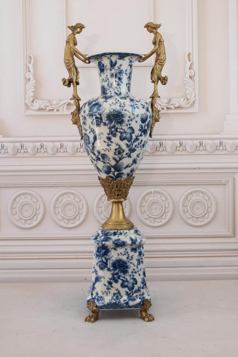 May include: A large, white porcelain vase with a blue floral pattern and gold accents. The vase has two gold handles in the shape of women holding up the vase. The vase is on a square base with gold feet.