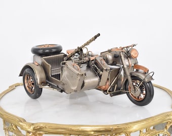 May include: A metal model of a vintage motorcycle with a sidecar, featuring a mounted machine gun, a spare tyre on the rear, and copper-toned wheel spokes. The vehicle is finished in a muted metallic grey with visible mechanical details.