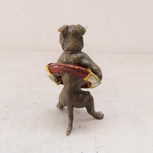 May include: A bronze-coloured dog figurine, viewed from the back, holding a red and gold object. The dog has a red collar. The figurine is on a white surface.