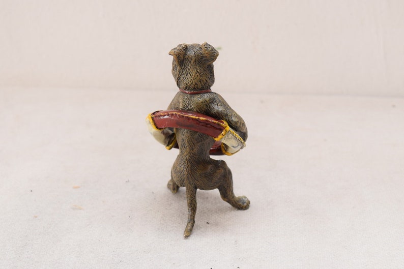 May include: A bronze-coloured dog figurine, viewed from the back, holding a red and gold object. The dog has a red collar. The figurine is on a white surface.