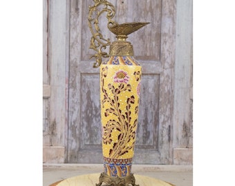 May include: A tall, yellow ceramic vase with a floral design and gold accents. The vase has a decorative brass spout and handle.