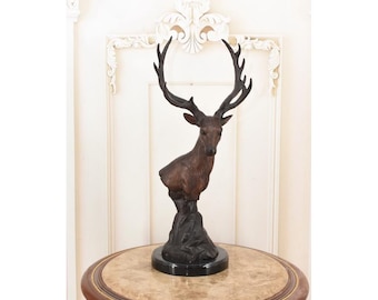 May include: A bronze-colored deer bust sculpture with large antlers. The deer is mounted on a black base that sits atop a round, marble-topped table. The background features a white wall with decorative molding.